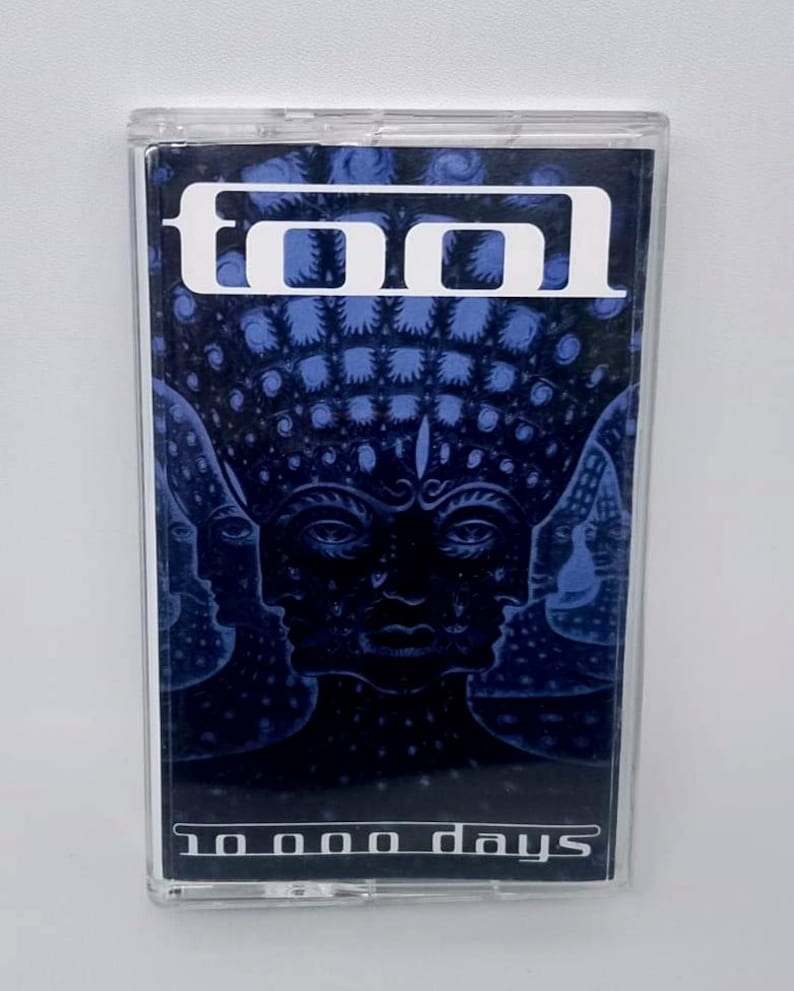 May include: A Tool cassette tape with a blue and black album cover. The cover features the band's logo in white at the top and the album title "10,000 Days" at the bottom. The artwork depicts multiple faces.