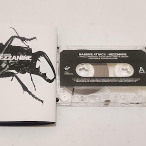 May include: A cassette tape of the album "Mezzanine" by Massive Attack. The cassette case is clear, and the album art features a black and white illustration of an insect. The album title and tracklist are visible.