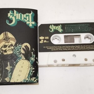 Op de afbeelding: Een witte cassetteband en een zwarte cassettehoes. De hoes toont een skeletfiguur met een pauselijke hoed die een tablet vasthoudt. De band heeft de bandnaam "Ghost" en de albumtitel "13 Commandments".