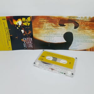 May include: A clear cassette tape with a yellow label and the text "1" is next to the open album cover of the Wu-Tang Clan's "Enter the Wu-Tang (36 Chambers)" album. The album cover is yellow and features the Wu-Tang Clan logo and album title.