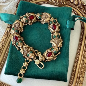 Può includere: Bracciale dorato con ciondoli a forma di rana con dettagli rossi e verdi. Il bracciale è presentato su un sacchetto di velluto verde, con una cornice dorata sullo sfondo. Chiusura a moschettone.