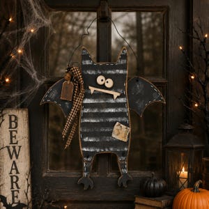 May include: A hanging bat decoration featuring a black and white striped body, two large round eyes, and two white fangs. The bat has black wings with white stitched details, a brown tag labeled "BOO" with a plaid ribbon, and a small patch labeled "31".