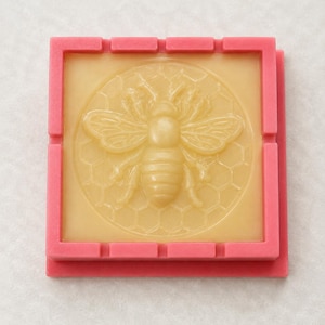 Può includere: Uno stampo in silicone quadrato di colore rosa vivace con un design circolare centrale. Il design raffigura una figura di ape in rilievo centrata su un dettagliato motivo a nido d'ape esagonale. Lo stampo è color beige chiaro e liscio.