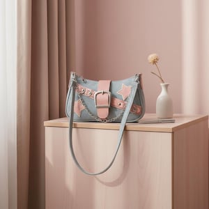 Puede incluir: Un bolso gris con detalles en rosa, incluyendo una hebilla, parches en forma de estrella y una correa. El bolso presenta una cadena plateada y una correa gris a juego. El bolso se exhibe sobre una superficie de madera clara.