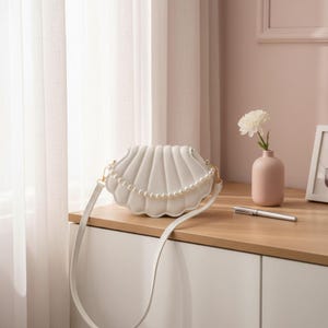 Könnte beinhalten: Eine weiße, muschelförmige Umhängetasche mit Perlenbesatz. Die Tasche hat einen langen, dünnen Riemen und goldfarbene Beschläge. Die Tasche liegt auf einer Holzoberfläche neben einer rosa Vase mit einer weißen Blume.