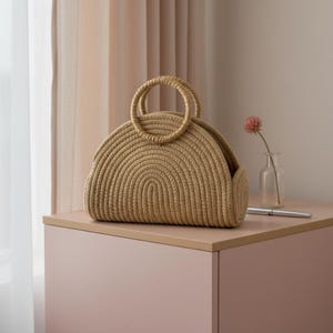 May include: A woven, tan-colored handbag with two circular handles. The bag has a rounded shape and a textured surface. It sits on a pink surface next to a small vase with a pink flower and a silver pen.