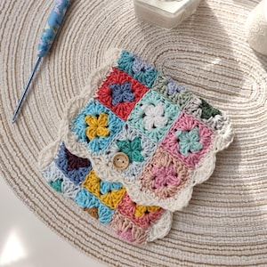 May include: A handmade crochet coaster with a button fastening, featuring a patchwork design in various colours and floral patterns. A blue crochet hook and a lit candle in a glass jar are also visible.