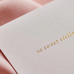 May include: A close-up of a white card with the gold text "xo sweet stella". The card is set against a backdrop of a soft, pink fabric. The card has a textured surface.