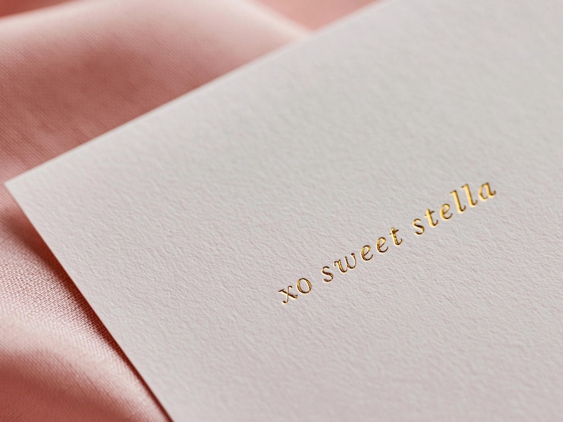 May include: A close-up of a white card with the gold text "xo sweet stella". The card is set against a backdrop of a soft, pink fabric. The card has a textured surface.