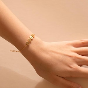 Op de afbeelding: Een goudkleurige armband met vier ronde bedels, elk met een enkele letter. De armband zit aan een fijne ketting en wordt om de pols gedragen. De letters vormen een woord of initialen. Neutrale achtergrond.