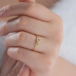 May include: A delicate gold ring featuring a small, dark gemstone and the letter "D". The ring is simple and elegant, with a thin band. The ring is worn on a finger.