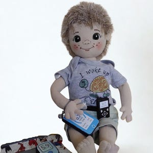 May include: A handmade doll wearing a grey t-shirt with a turtle graphic and the text "I woke up". The doll is holding a blue device and a black device. A case with a blood glucose meter is in front of the doll.