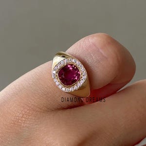 Könnte beinhalten: Ein goldener Ring mit einem großen, oval geschliffenen, tiefrosa Edelstein, umgeben von einem Kranz aus kleinen, klaren Edelsteinen. Der Ring hat einen glatten, polierten Schaft und der Text "DIAMOND DREAMS" ist sichtbar.