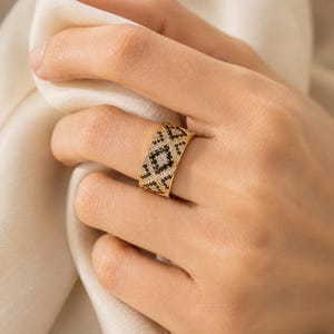 May include: A wide gold band ring featuring a geometric pattern of alternating black and clear stones. The design consists of small triangular and diamond-shaped sections filled with pav&eacute;-set crystals, creating a textured, high-contrast appearance.