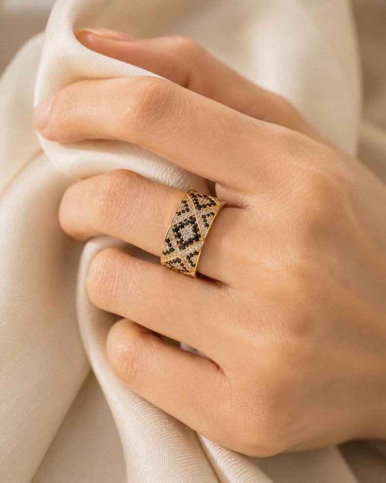 May include: A wide gold band ring featuring a geometric pattern of alternating black and clear stones. The design consists of small triangular and diamond-shaped sections filled with pav&eacute;-set crystals, creating a textured, high-contrast appearance.
