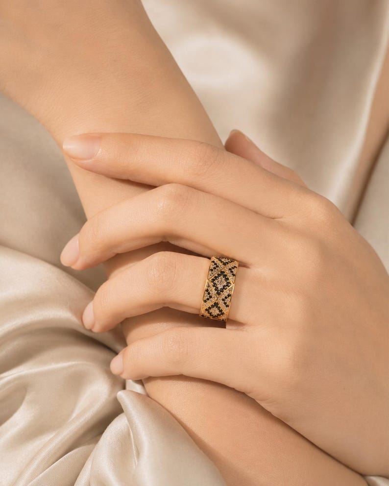 May include: A wide gold band ring featuring a repeating geometric diamond pattern. The design is encrusted with small, alternating black and clear stones that create a textured, high-contrast look across the entire surface of the metal band.