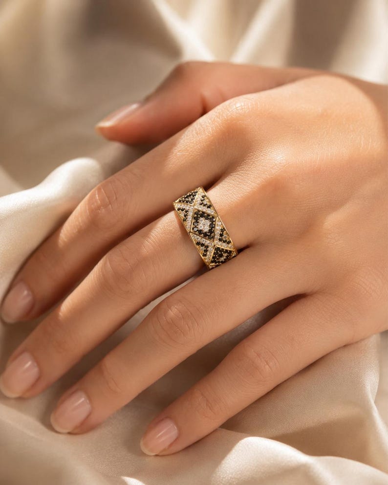 May include: A wide gold band ring featuring a geometric pattern of alternating triangles and diamonds. The design is encrusted with small, sparkling clear white stones and contrasting black stones, creating a detailed, textured surface across the band.