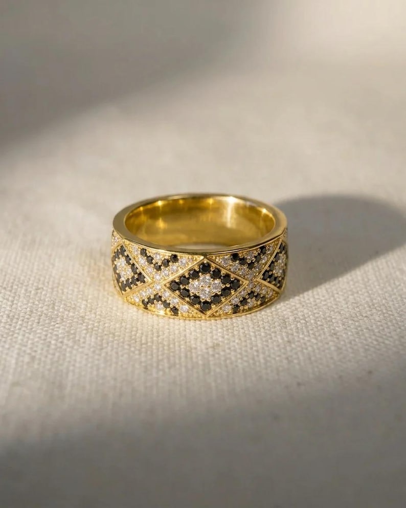 May include: A wide gold band ring featuring a geometric diamond pattern across the outer surface. The design is encrusted with alternating small clear white stones and small black stones, creating a high-contrast, repeating lattice motif.