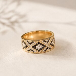 May include: A wide gold band ring featuring a geometric diamond pattern across the surface. The design is encrusted with alternating small round black and clear white stones set within the metal, creating a repeating lattice motif throughout.