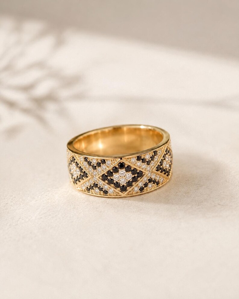 May include: A wide gold band ring featuring a geometric diamond pattern across the surface. The design is encrusted with alternating small round black and clear white stones set within the metal, creating a repeating lattice motif throughout.