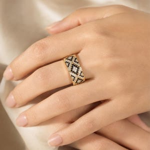 May include: A wide gold band ring featuring a geometric pattern of alternating black and white stones. The design consists of repeating diamond shapes filled with small, sparkling black and clear stones set into the polished gold metal surface.