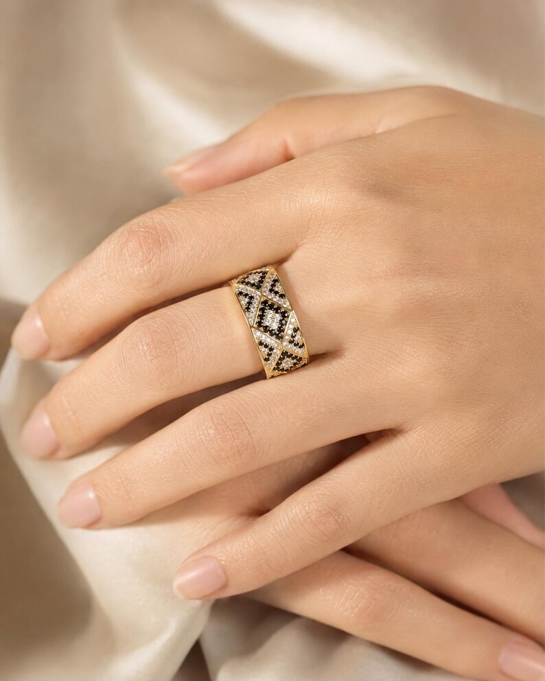 May include: A wide gold band ring featuring a geometric pattern of alternating black and white stones. The design consists of repeating diamond shapes filled with small, sparkling black and clear stones set into the polished gold metal surface.