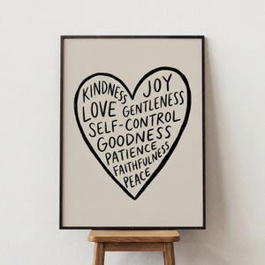 Puede incluir: Un cuadro enmarcado con un corazón negro lleno de palabras como "Kindness", "Love" y "Joy". El corazón está delineado en negro sobre un fondo crema, dentro de un marco negro. Descansa sobre un pequeño taburete de madera.