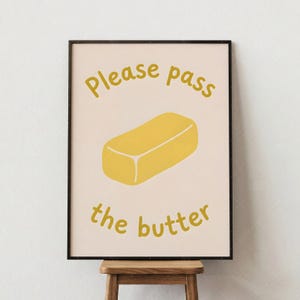 May include: A framed print featuring a yellow illustration of a butter stick on a light pink background. The words "Please pass the butter" are written in yellow above and below the butter illustration. The frame is black.