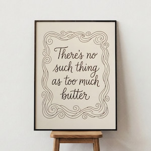 Puede incluir: Un cuadro enmarcado con la frase "There's no such thing as too much butter" en una elegante escritura. El texto está en un marco decorativo con líneas onduladas, sobre un fondo crema. El marco es negro.