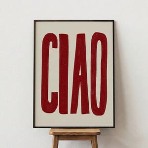 Op de afbeelding: Een ingelijste kunstdruk met het Italiaanse woord "CIAO" in dikke, rode letters tegen een crèmekleurige achtergrond. Het kunstwerk wordt gepresenteerd in een zwarte lijst en staat op een kleine houten kruk.