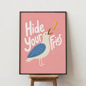 May include: A framed art print featuring a cartoon seagull with a blue wing, eating a french fry. The pink background has the words "Hide Your Fries" in white script. The print is in a black frame.