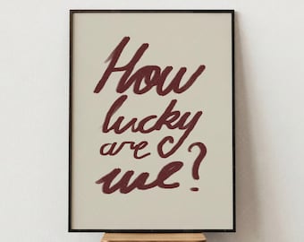May include: A framed print with the text "How lucky are we?" in a handwritten style, burgundy color. The print is set against a cream background and framed in black. The artwork is displayed on a small wooden stool.
