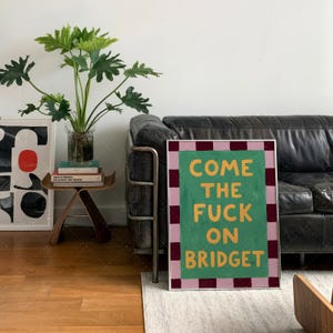 Come The Fuck On Bridget bold typography print in frame on wall near leather sofa, retro poster mockup in contemporary living room with mid-century furniture and plants