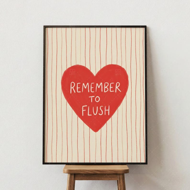 Remember To Flush bathroom wall art print, product detail closeup showing red heart with humorous typography on cream background with vertical red stripes, displayed in black frame on wooden stool against neutral wall
