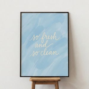 Könnte beinhalten: Gerahmter Druck mit hellblauem Aquarellhintergrund und den Worten "so fresh and so clean" in eleganter Schreibschrift. Der Rahmen ist schwarz, und der Druck wird auf einem kleinen Holzschemel präsentiert.