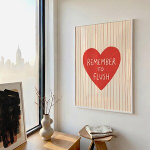 Bathroom humor wall art in frame on wall by window, Remember To Flush red heart poster mounted in bright room with city view, wooden console table and white vase, showing contemporary interior styling