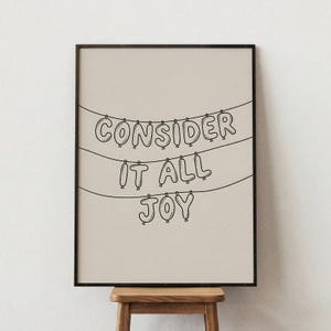 以下が含まれることがあります： 「CONSIDER IT ALL JOY」の文字が黒い風船のような文字で書かれたフレーム入りのプリント。ベージュの背景に3本の水平線から文字が吊り下げられています。フレームは濃い茶色で、木製のスツールの上にプリントが飾られています。