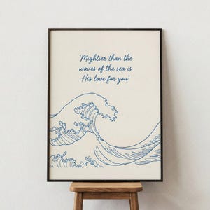 Könnte beinhalten: Gerahmter Kunstdruck mit cremefarbenem Hintergrund, der eine blaue Strichzeichnung einer Welle und das Zitat "Mächtiger als die Wellen des Meeres ist Seine Liebe zu dir" zeigt. Das Kunstwerk befindet sich in einem schwarzen Rahmen und steht auf einem Holzschemel.