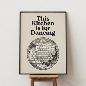 Puede incluir: Un estampado enmarcado con el texto "This Kitchen is for Dancing" sobre una ilustración de bola de discoteca. El estampado tiene un fondo crema y un marco negro. La bola de discoteca está hecha de pequeños cuadrados reflectantes.