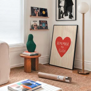 Humorous bathroom wall art in frame on wall, Remember To Flush red heart print shown in contemporary room setting with wooden shelves, copper lamp, and modern furnishings, demonstrating stylish home decor application
