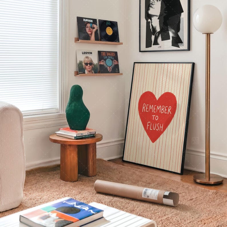 Humorous bathroom wall art in frame on wall, Remember To Flush red heart print shown in contemporary room setting with wooden shelves, copper lamp, and modern furnishings, demonstrating stylish home decor application