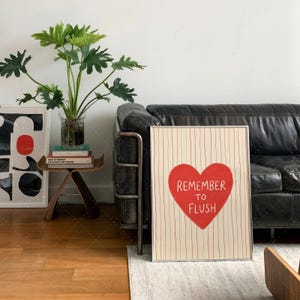 Remember To Flush typography print in frame on wall near black leather sofa, funny bathroom art poster with red heart design displayed in mid-century modern living room with houseplant and wooden side table