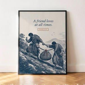 Puede incluir: Una impresión artística enmarcada con una ilustración en tonos azules de dos figuras con túnicas subiendo un sendero de montaña rocoso mientras cargan un saco grande en un palo. El texto dice A friend loves at all times. Proverbs 17:17.