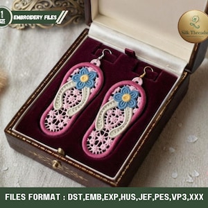 Könnte beinhalten: Ein Paar rosa-weiße, gestickte Flip-Flop-Ohrringe mit blauen und gelben Blumenakzenten, präsentiert in einer braunen Schmuckschatulle mit bordeauxrotem Innenraum. Die Ohrringe sind mit filigranen Spitzenmustern und goldfarbenen Haken versehen.