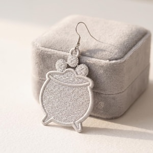 May include: A light grey embroidered earring shaped like a cauldron with three small circular bubbles rising from the top. The design features a stitched texture throughout the surface and is attached to a silver-toned metal fishhook earring wire.
