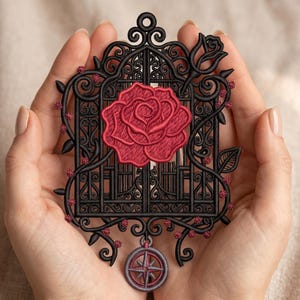 May include: An embroidered design featuring a black wrought iron gate with a large red rose at the center. The gate is adorned with vines and small red flowers. A compass charm hangs below. The design is held in two hands.