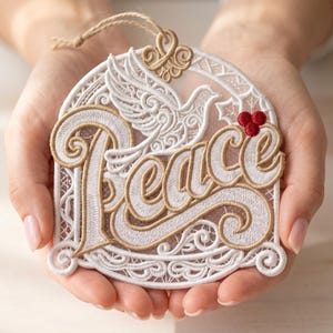 May include: Embroidered ornament with the word "Peace" in a decorative font, featuring a dove and red berries. The ornament has a white and gold color scheme with intricate lace-like detailing and a twine loop for hanging.