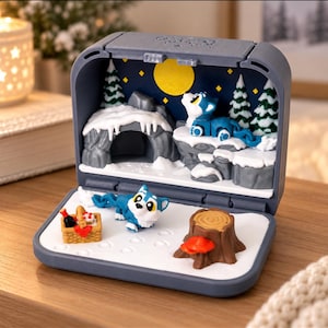 May include: A gray plastic clamshell case containing a miniature diorama. The top section features a blue wolf figurine on a snowy cliff under a yellow moon. The bottom section includes a blue wolf, a picnic basket, and a tree stump.
