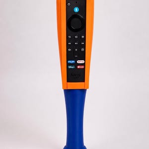 May include: A black Fire TV remote control encased in a bright orange protective sleeve, attached to a solid blue baseball bat-shaped handle. The remote features standard navigation buttons, a microphone icon, and logos for Prime Video, Netflix, Disney+, and YouTube.