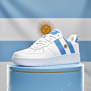 Puede incluir: Zapatilla de cuero blanca con rayas celeste y un emblema de sol, inspirada en la bandera argentina. El zapato presenta una suela y cordones blancos, sobre un fondo celeste con la bandera argentina.